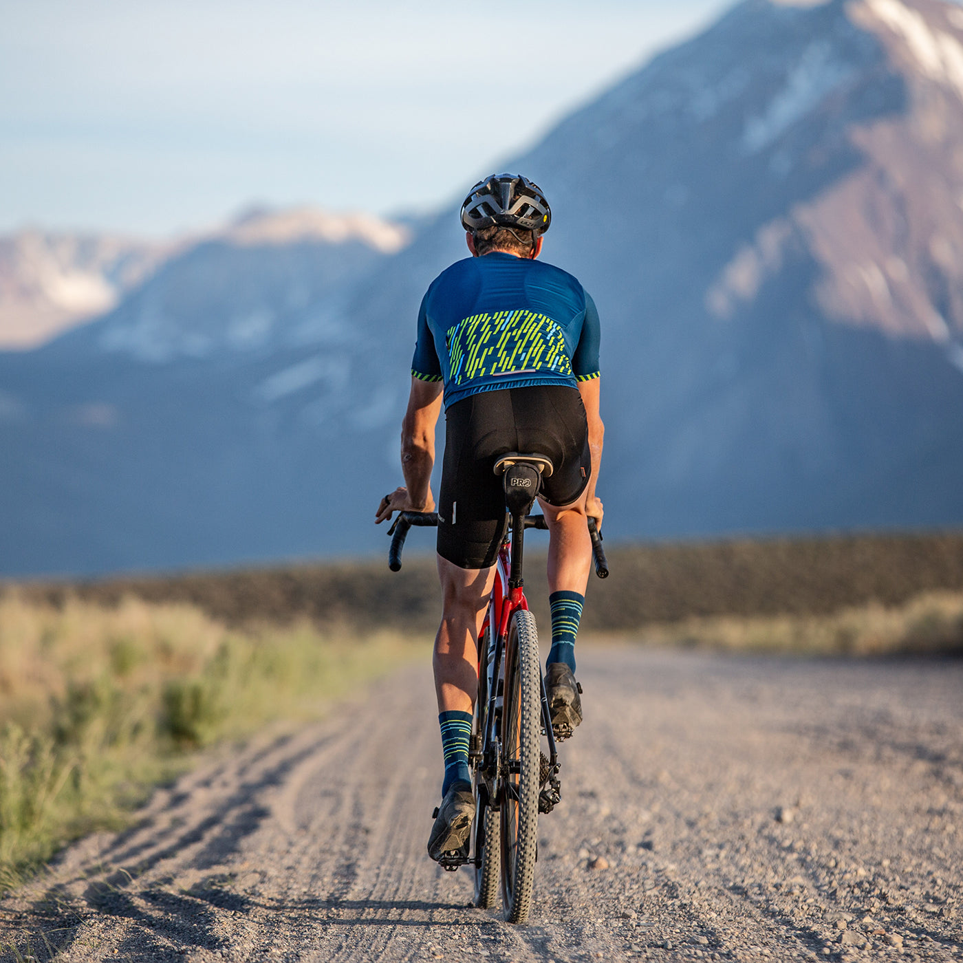 Solo cyclist riding down long gravel road in Revel jersey in baltic blue color